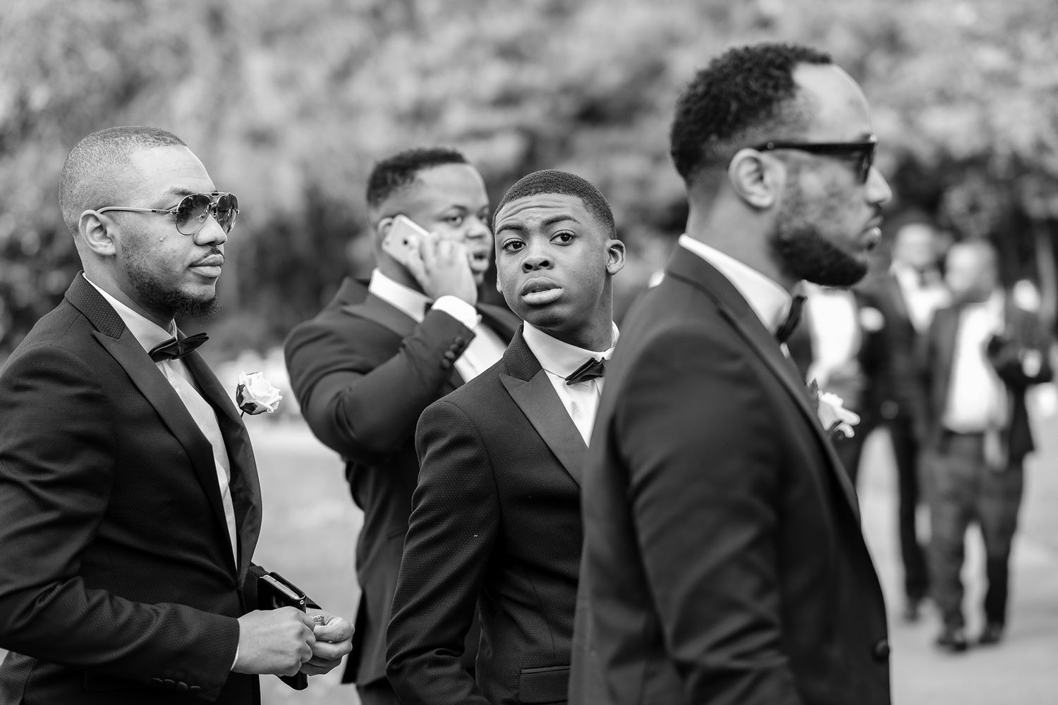 Groomsmen in black suits and sunglasses gather outside during wedding celebrations.