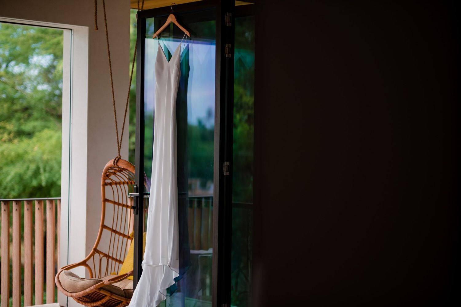 A white wedding dress hangs elegantly by a window next to a rattan chair.