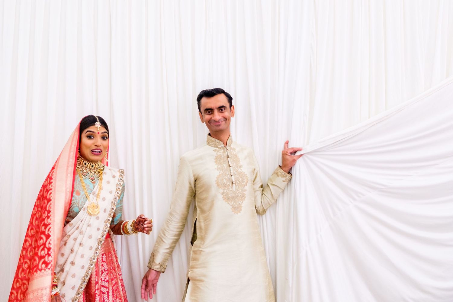 A couple in traditional South Asian wedding attire pose against a white backdrop.