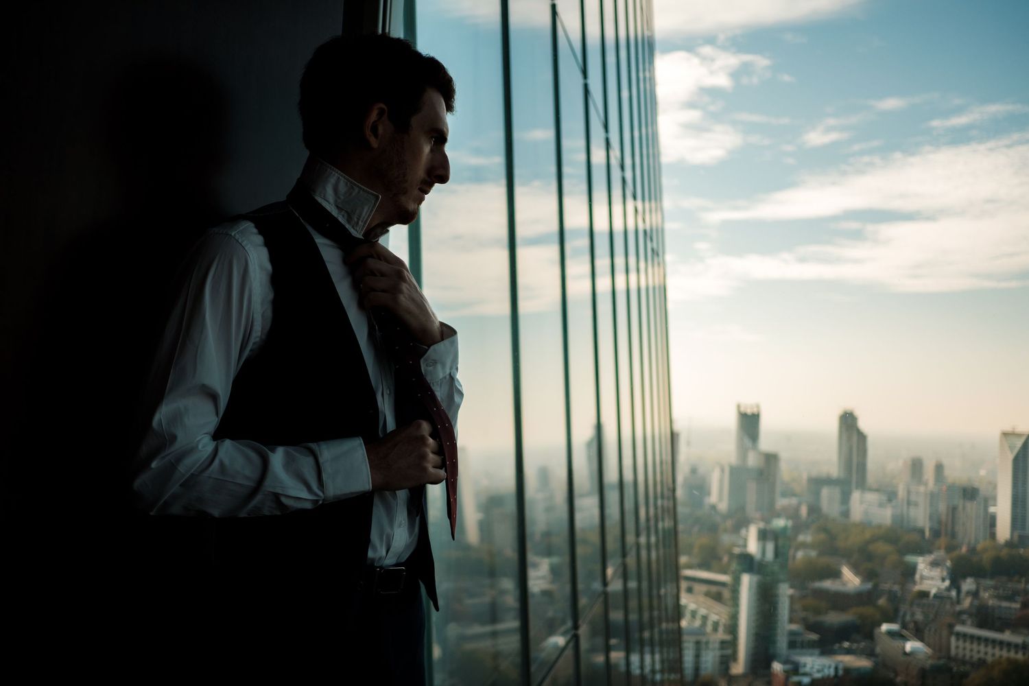 A silhouetted figure stands by floor-to-ceiling windows overlooking a cityscape at sunset with glass reflections.