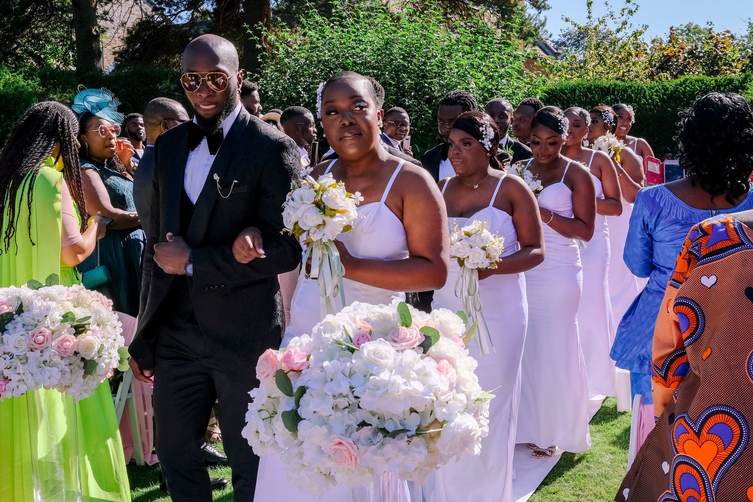 Wedding party in bright orange and white dresses poses outdoors in a garden setting during summer celebration.