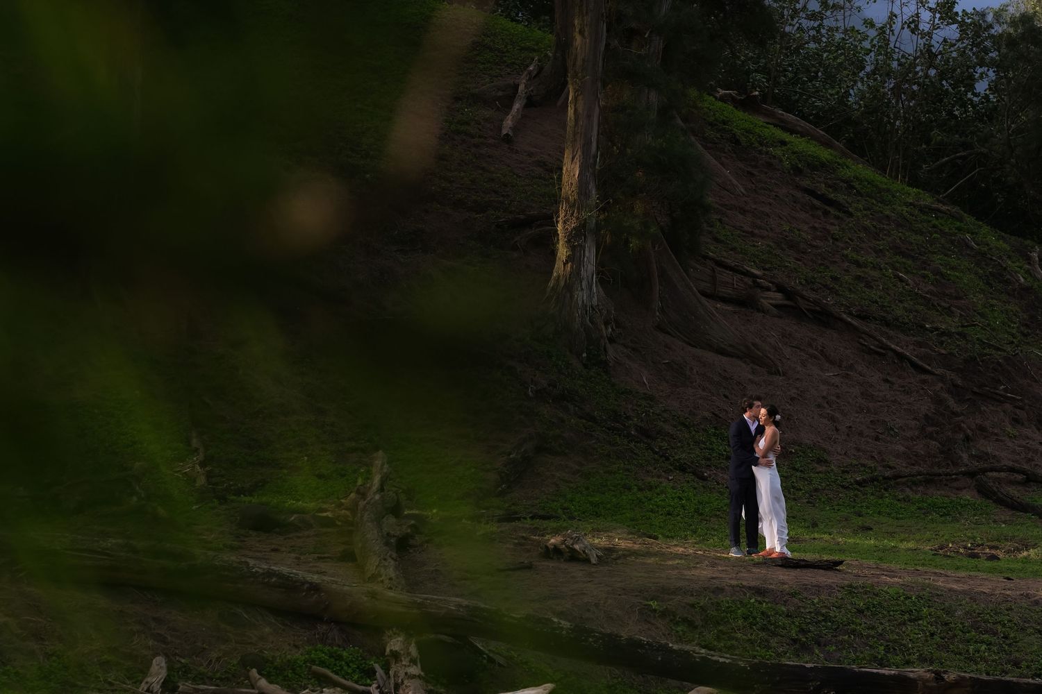 A couple in formal attire poses for wedding photos in front of a dramatic mountain landscape at sunset.