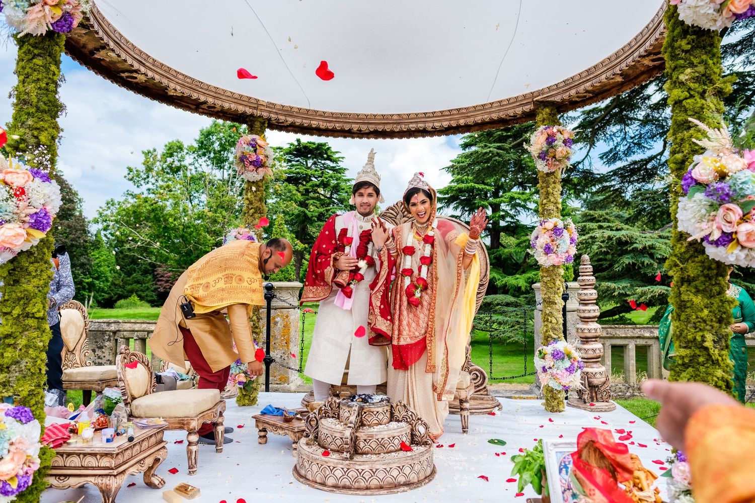 Ornate wedding ceremony under a circular mandap decorated with pink and white flowers in a garden setting.