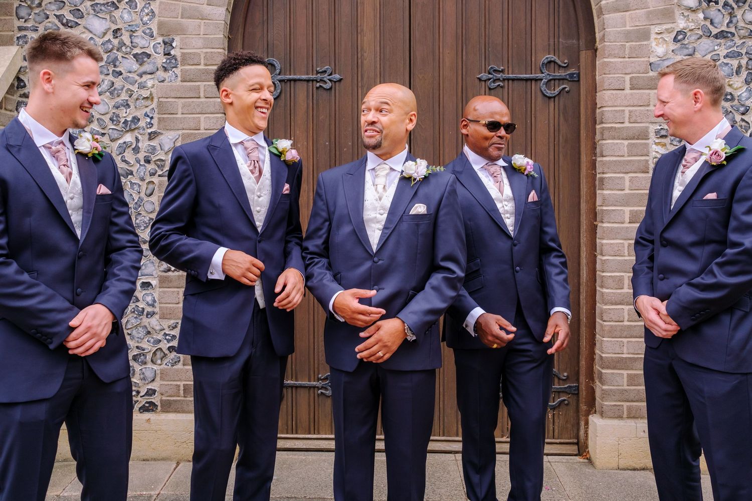Groomsmen in navy suits stand together laughing and chatting outside a brick church building.
