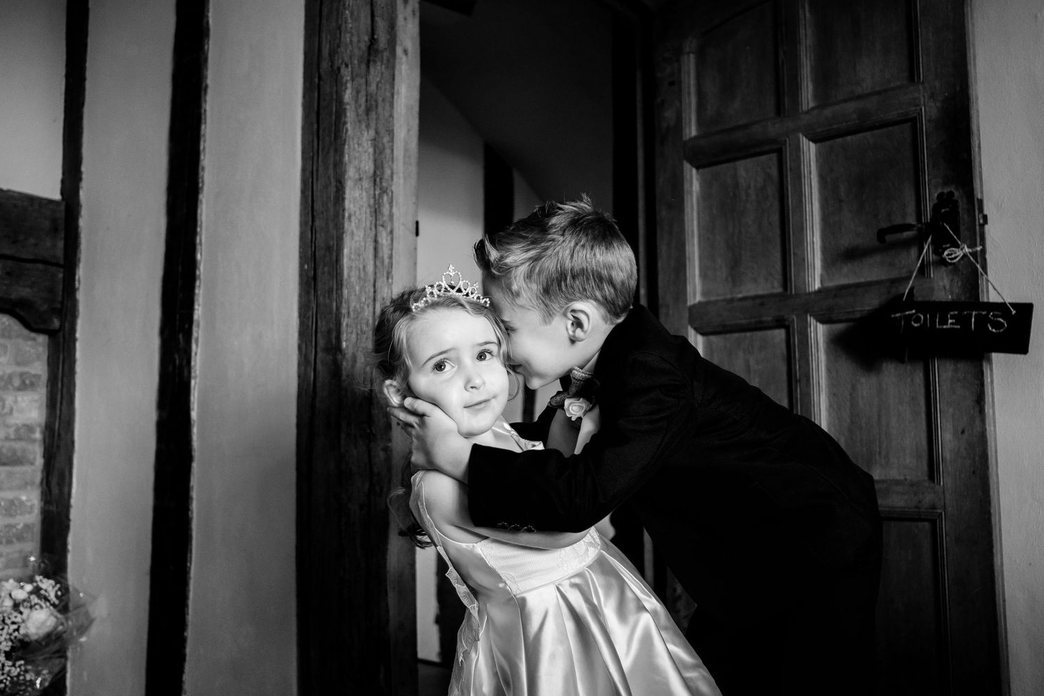 A vintage black and white photo capturing an intimate moment between two children at a doorway.