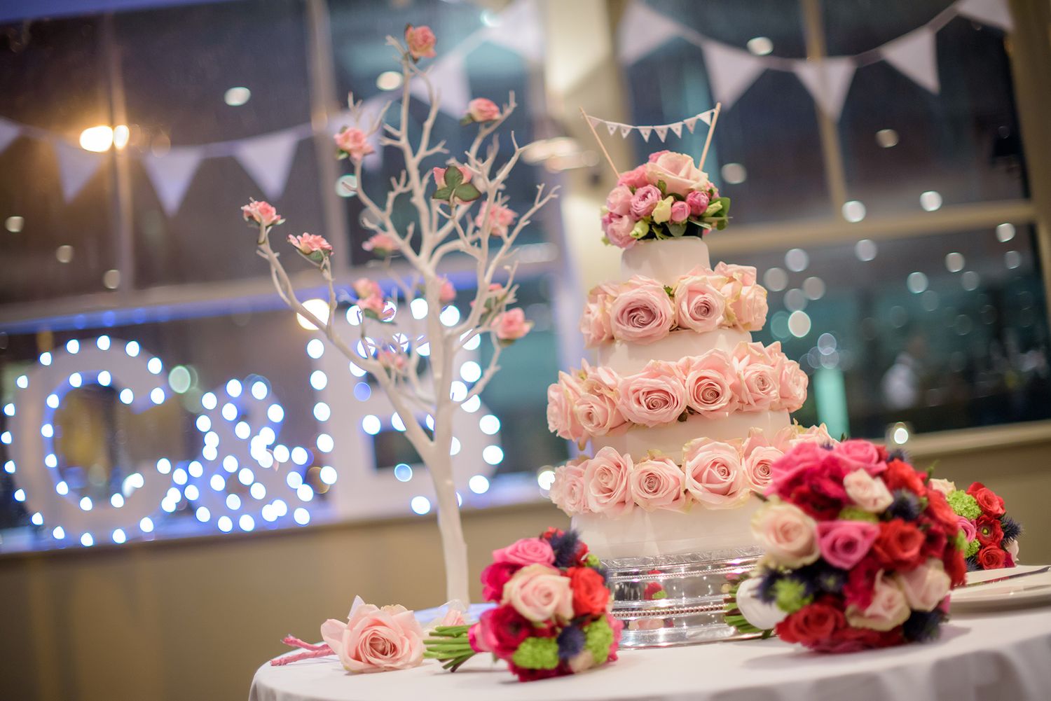 A wedding reception venue decorated with string lights and featuring a tiered pink floral cake centerpiece.