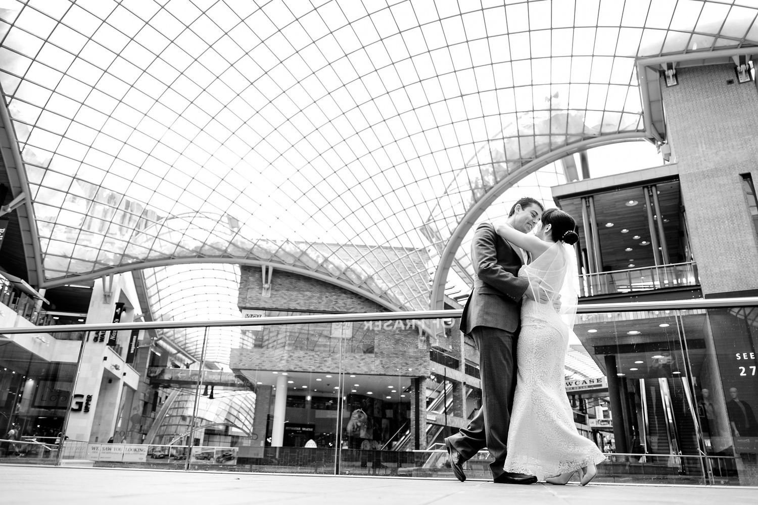 Wedding couple embracing under modern glass architecture in black and white photograph.