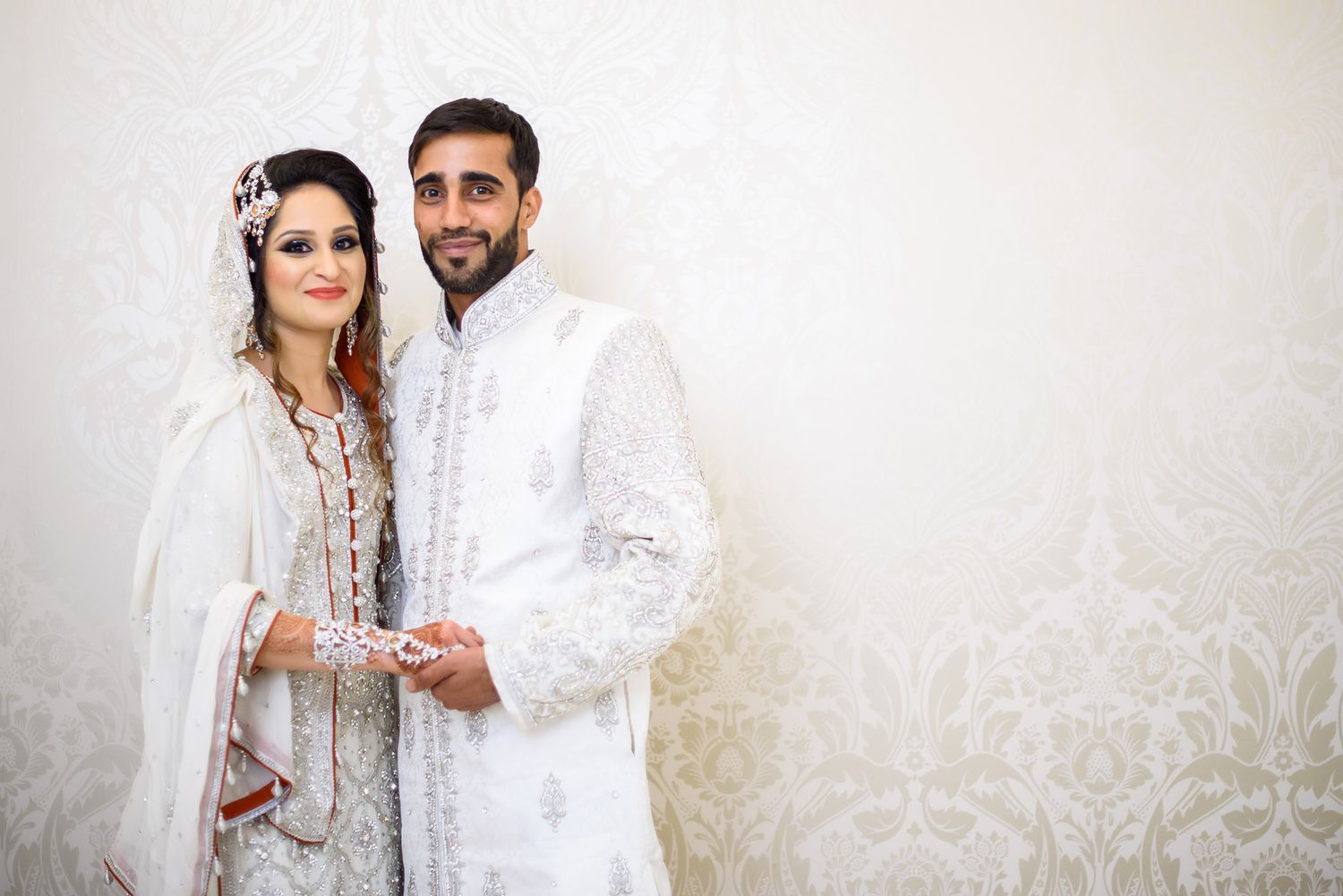 Wedding couple in white traditional outfits poses against a light patterned backdrop.