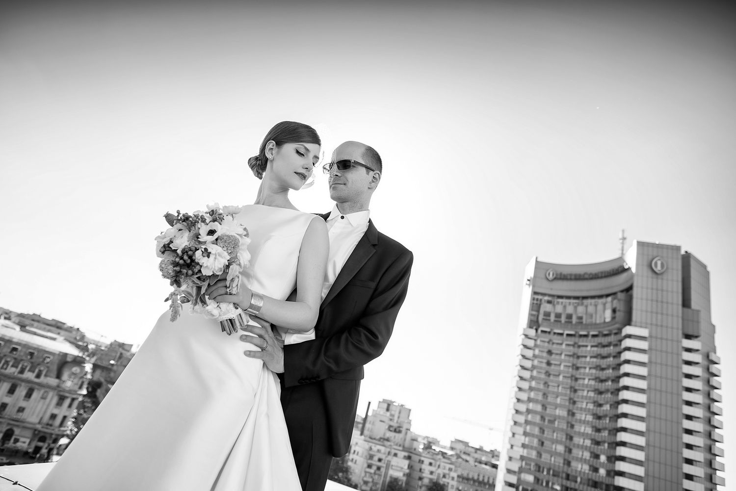 A couple in wedding attire share an intimate moment against a blue sky backdrop with city buildings.