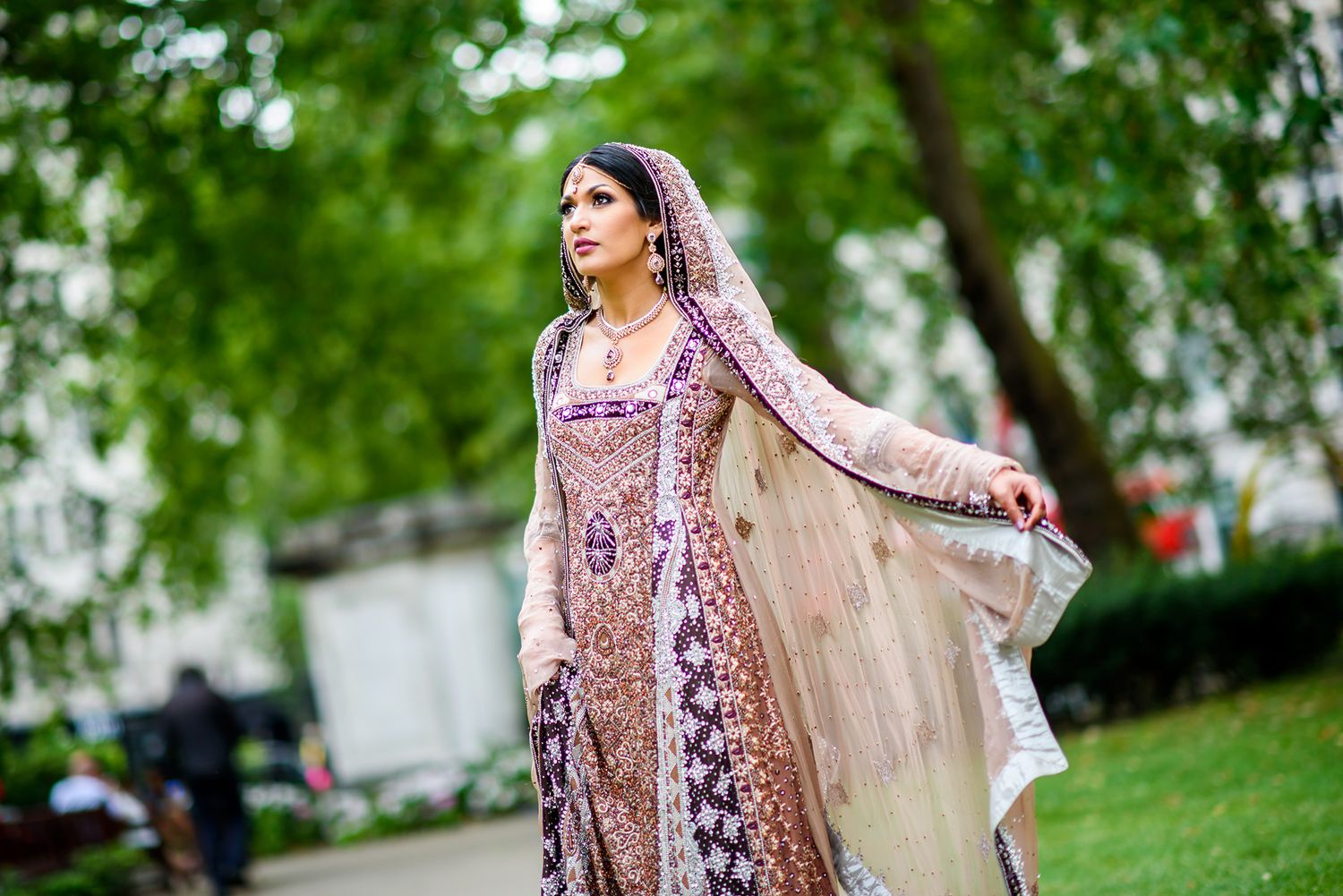 Bride in traditional South Asian attire with delicate embroidery and veil stands in a garden setting.