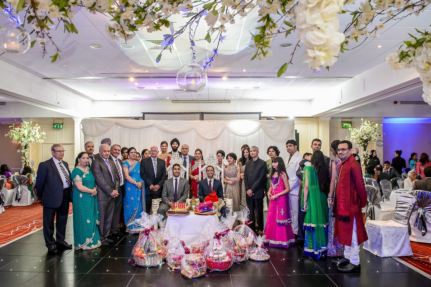 Large group photo of wedding guests in colorful traditional attire under floral ceiling decorations.