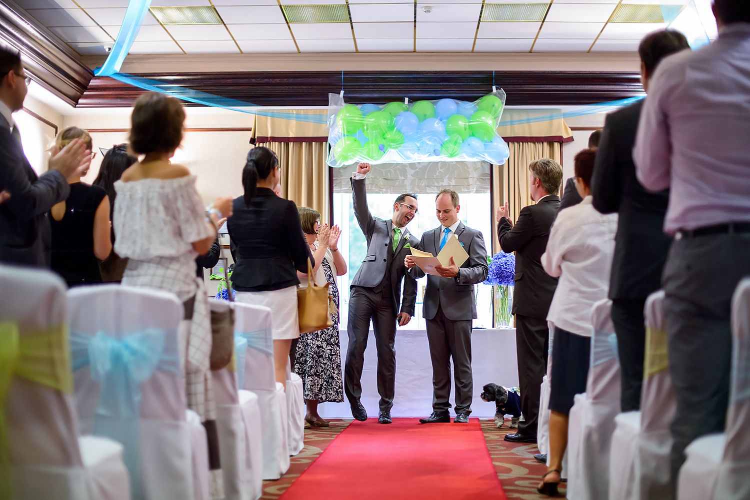 Wedding guests watch ceremony from white chairs along red carpet aisle under colorful lights.