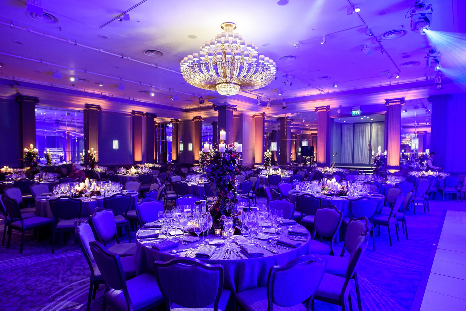 Grand ballroom bathed in purple lighting with crystal chandeliers and formal dining setup.