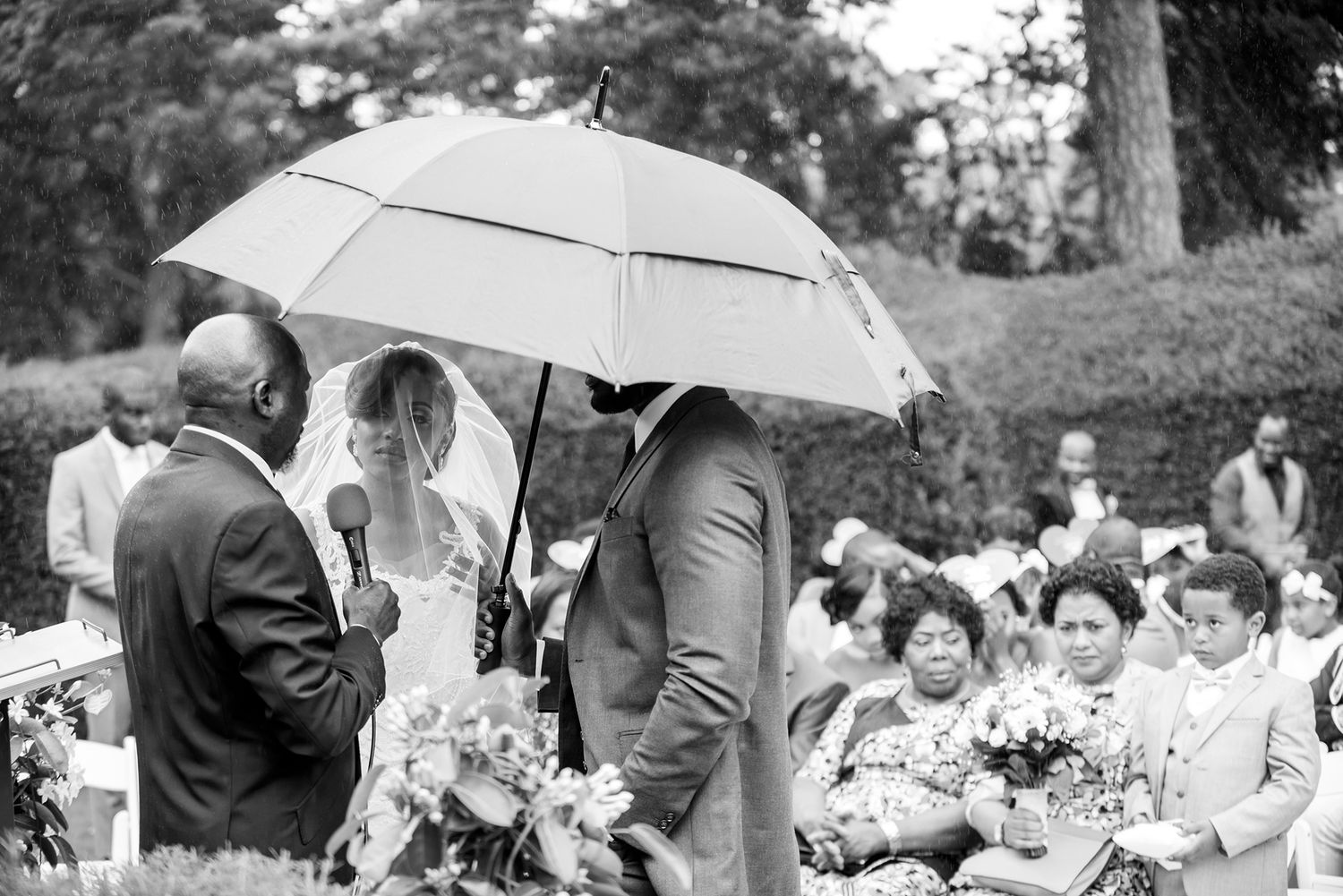 Wedding guests gather under white umbrellas during an outdoor garden ceremony.