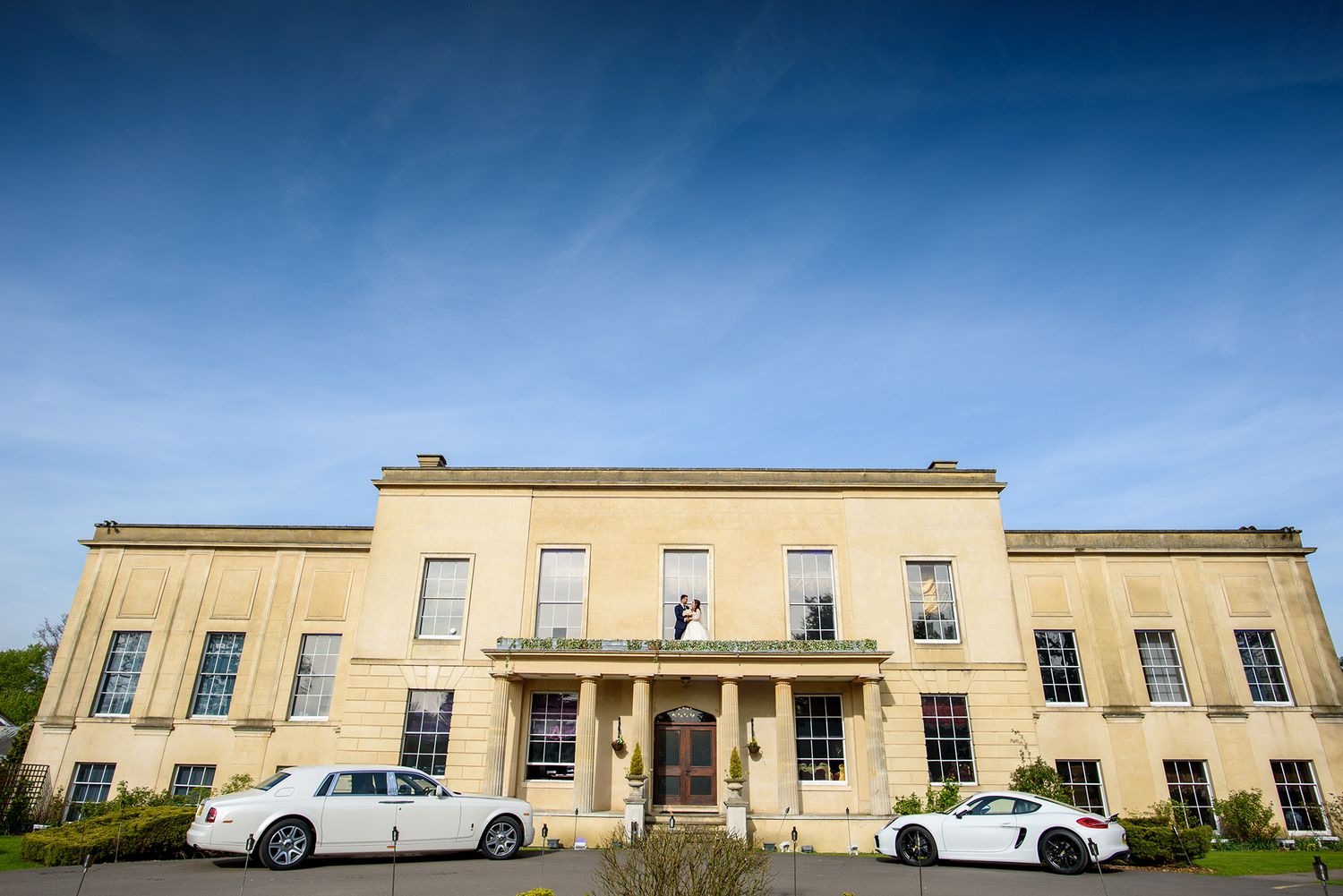 Long beige colored modern building with multiple windows and parked cars under blue sky.
