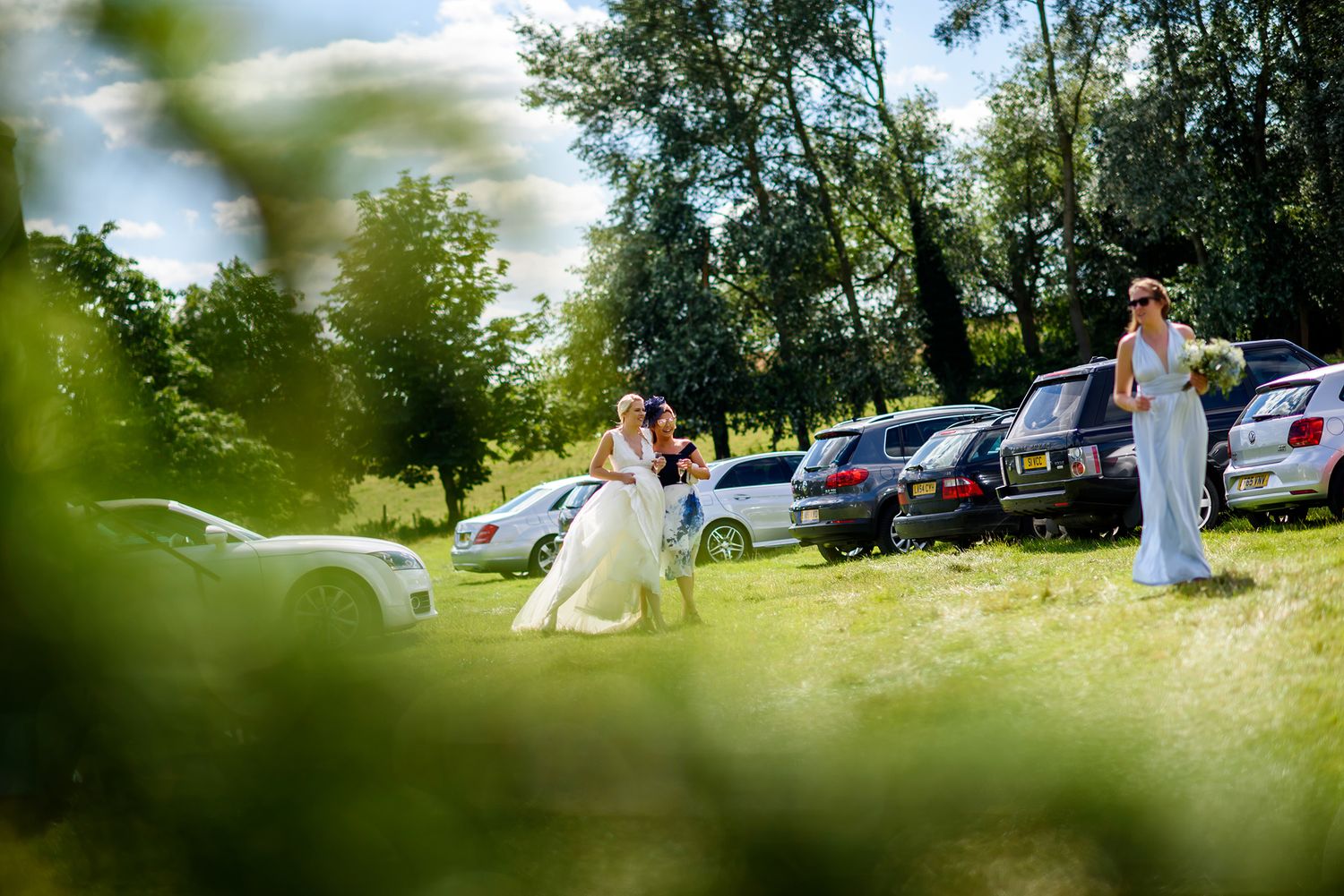 Distant view of wedding celebration through trees with parked cars visible.