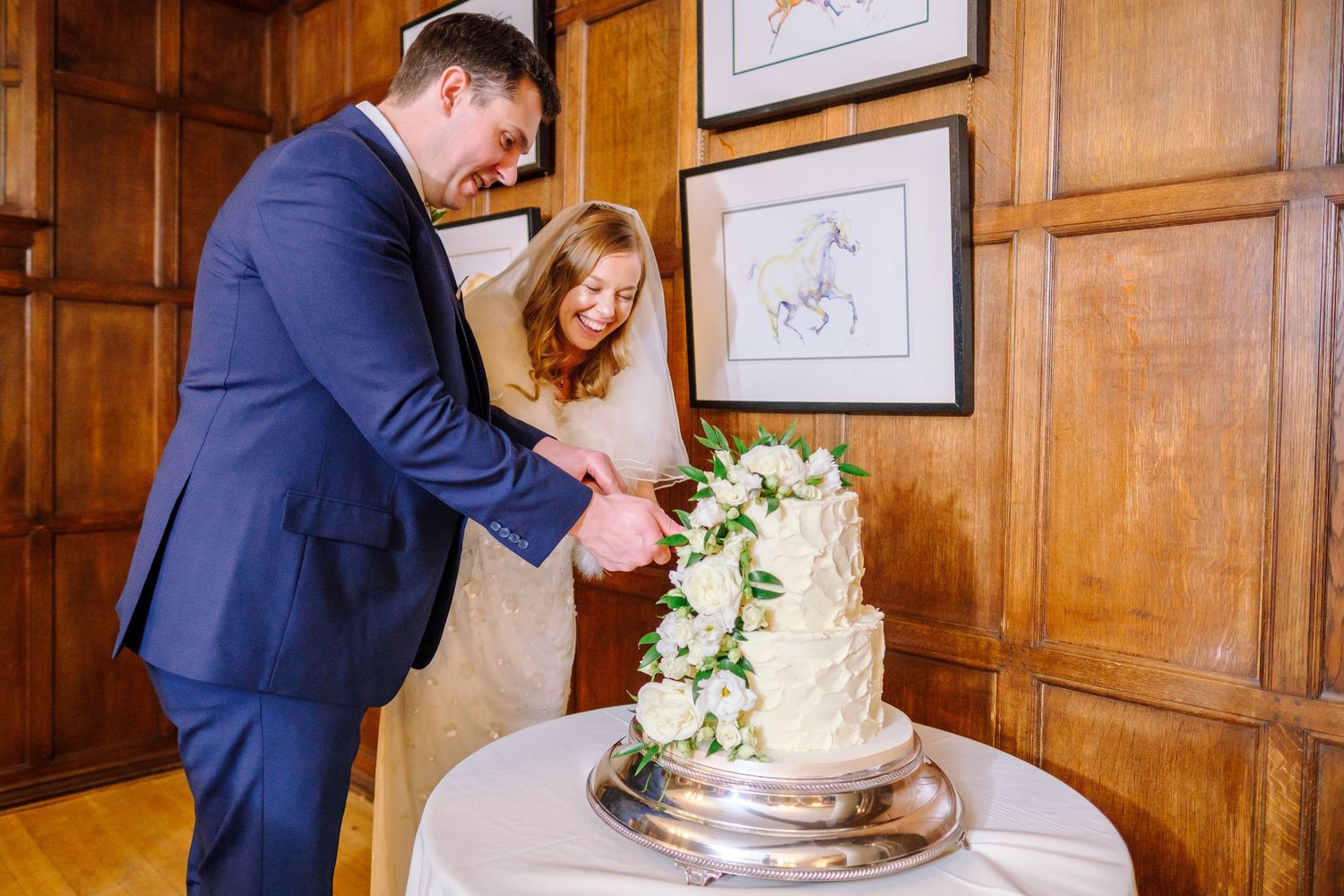 Couple cutting their elegant silver and white wedding cake in wood-paneled room.