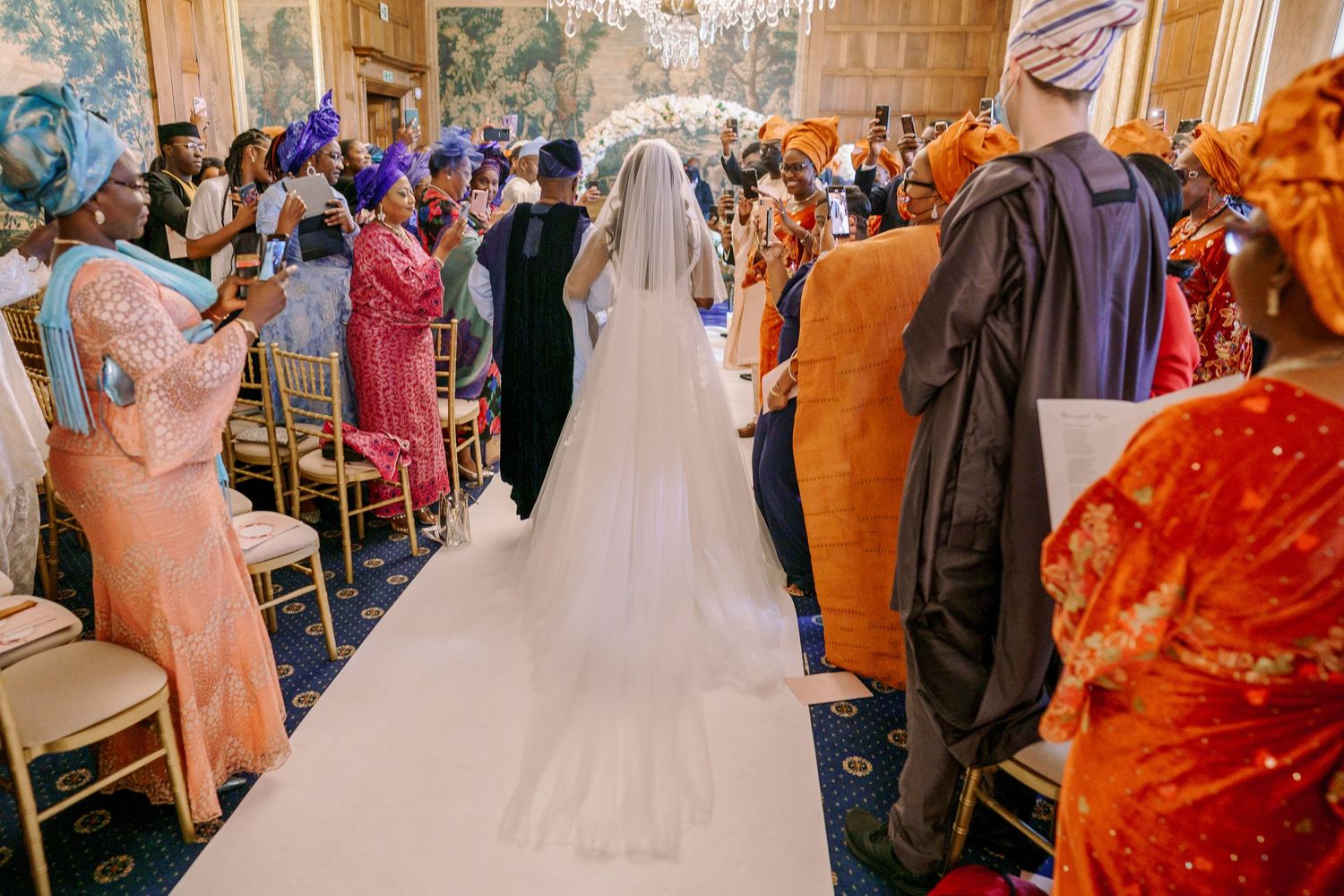 A bride in a flowing white gown walks down the aisle as wedding guests watch from their seats in an ornate venue.