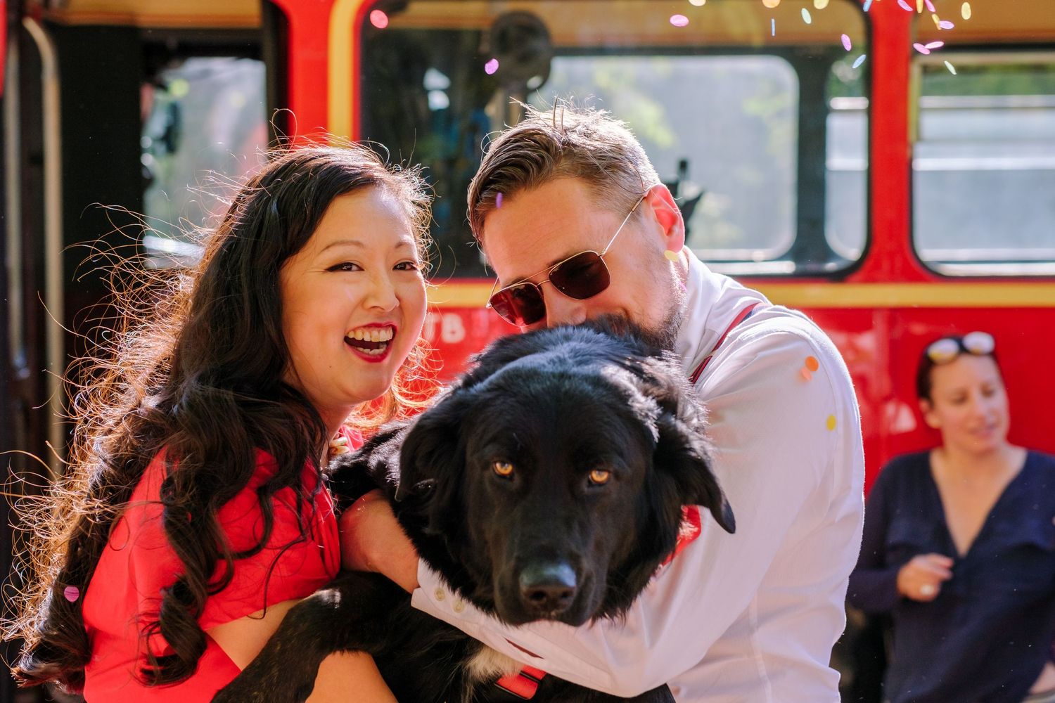 Black dog and couple share a playful moment in front of a red double-decker bus.