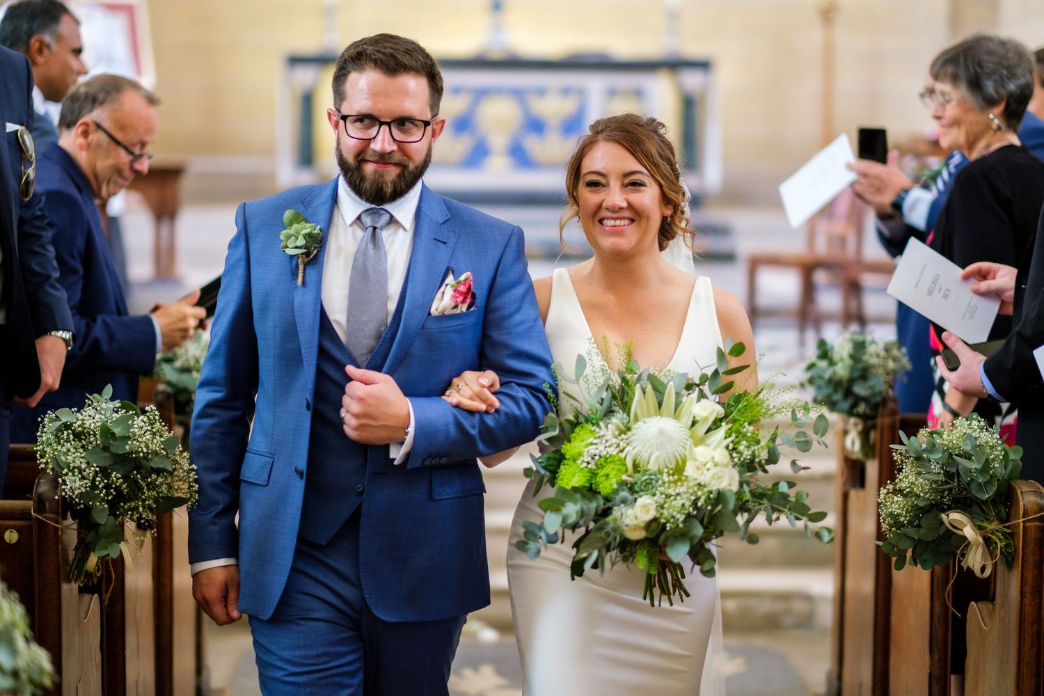 A joyful couple walks down the church aisle after their wedding ceremony, with the bride holding a white floral bouquet.