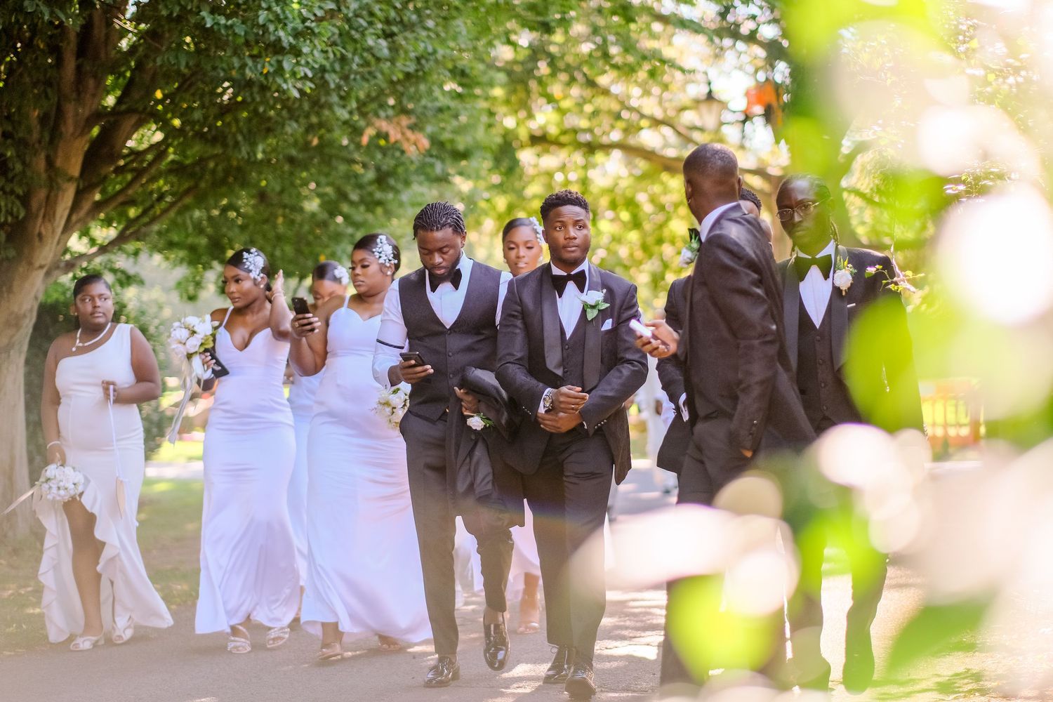 Wedding guests in white attire gather outdoors under green trees for a celebration.
