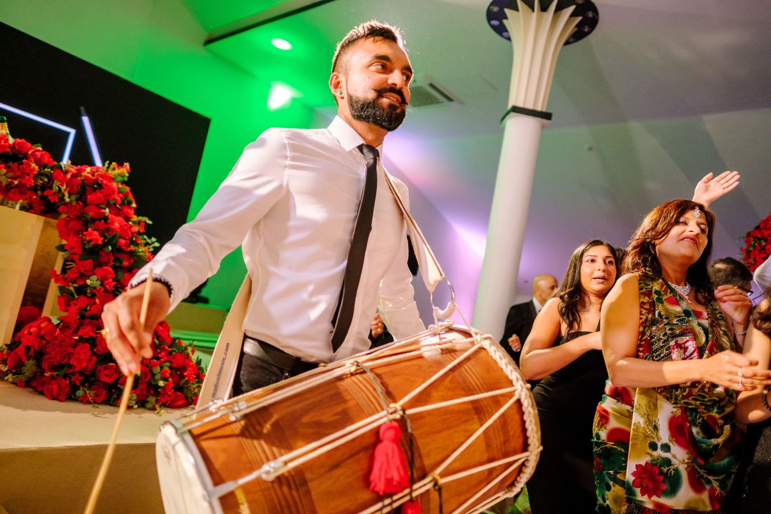 Energetic dhol drummer performs at wedding reception with green lighting and red floral decorations.
