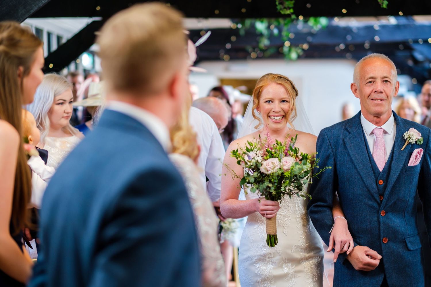 Wedding guests gather outdoors at a sunny venue with natural greenery.