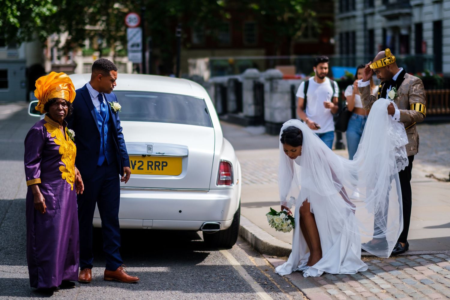 Wedding party members walk along city sidewalk with bride in white dress and bridesmaids in purple.
