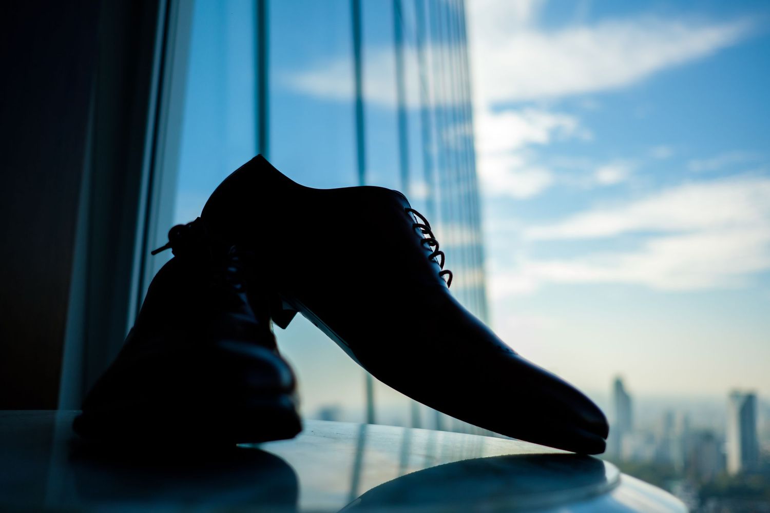 A series of silhouetted dress shoes against a bright window with a cityscape background and blue sky.