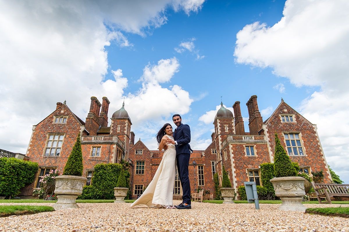 Wedding portrait in front of magnificent Tudor-style mansion under blue summer sky.