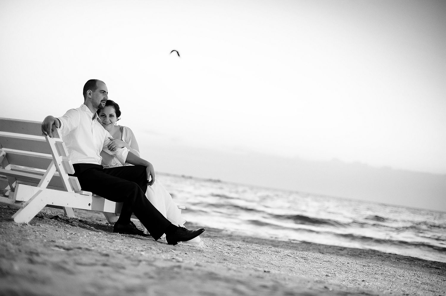 A black and white photo of a couple sitting on a beach chair near the ocean.