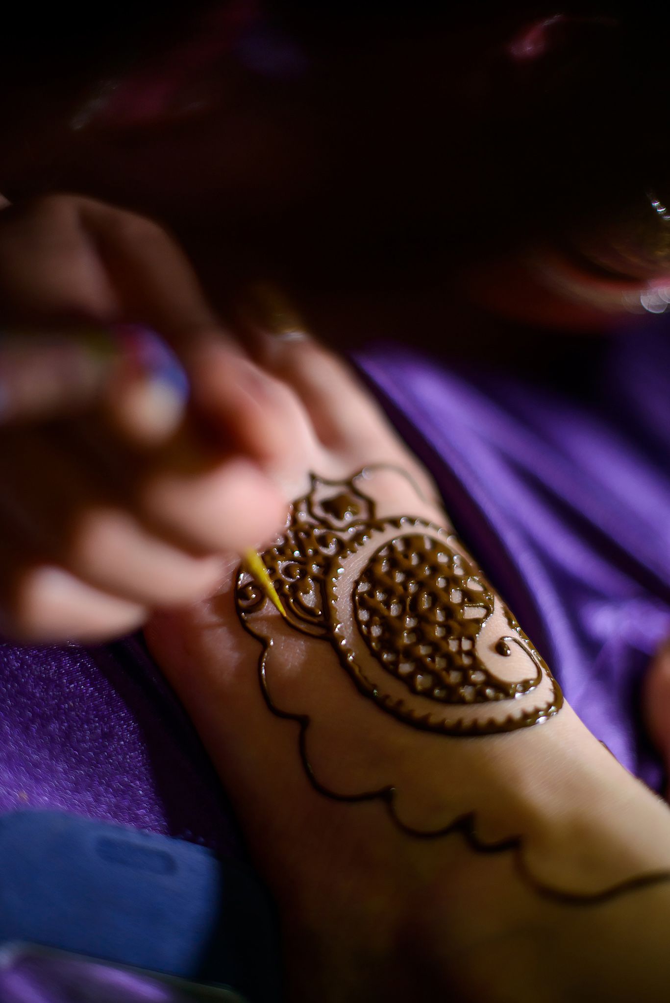 Detailed shot of intricate henna design being applied on hand against purple background.