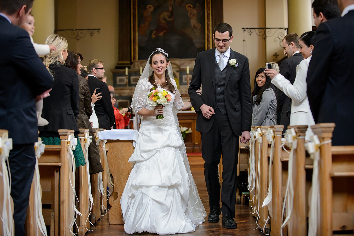 Wedding ceremony proceedings in church with guests seated in wooden pews.