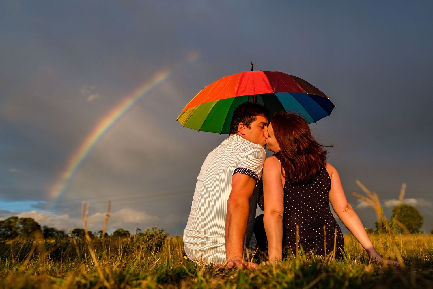 Rainbow arches over pastoral landscape with figures sitting together beneath colorful umbrellas at sunset.