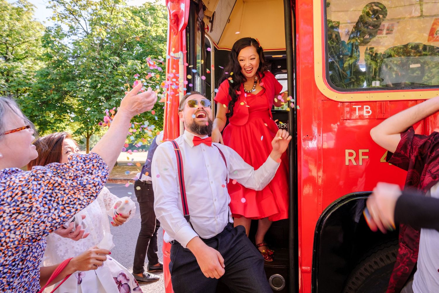 Wedding celebration scene outside a red London bus with confetti being thrown in the air.