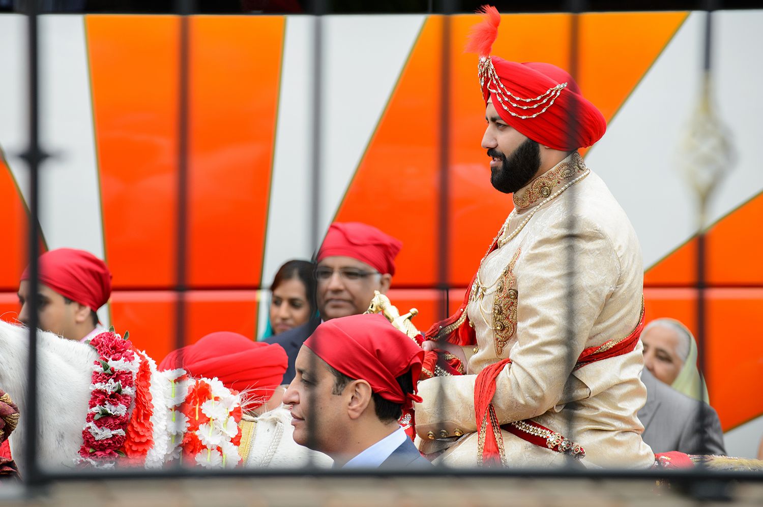 Wedding celebration with participants wearing traditional red turbans against an orange backdrop.