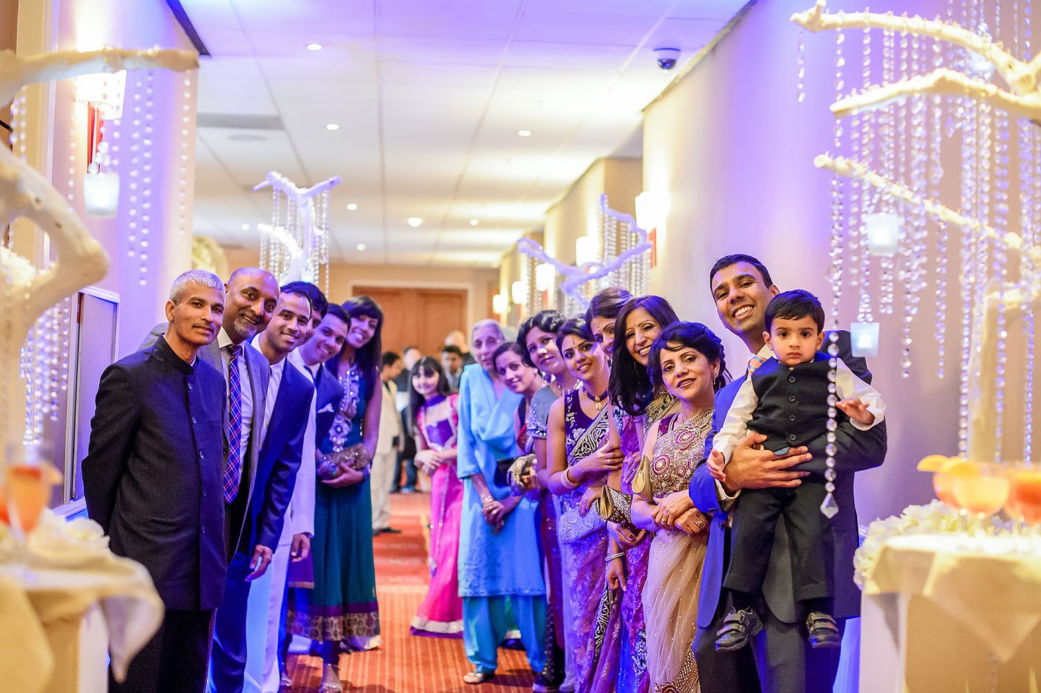 Wedding guests in colorful traditional attire gather in a hallway decorated with purple lights.