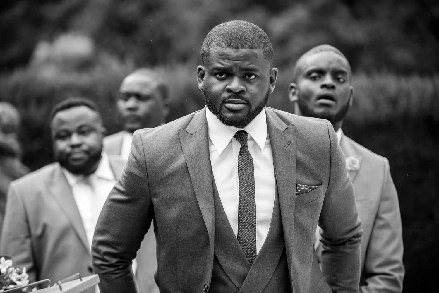 Groomsmen in dark suits pose together outdoors for formal wedding photos.