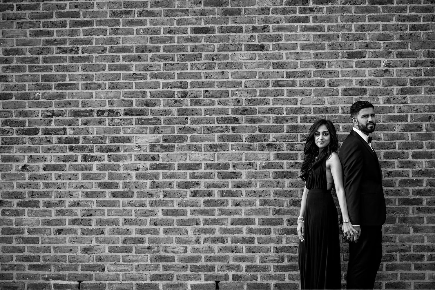 Stylish couple poses against red brick wall in dramatic black and white portrait.