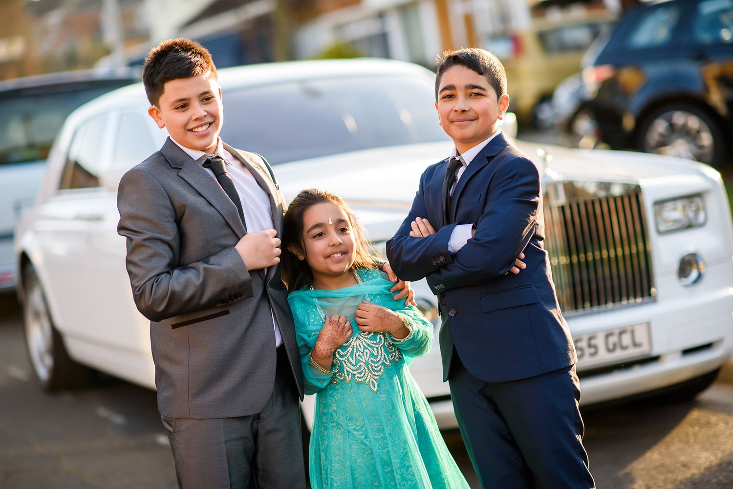 Three people stand together near a luxury white Rolls Royce at a formal event.