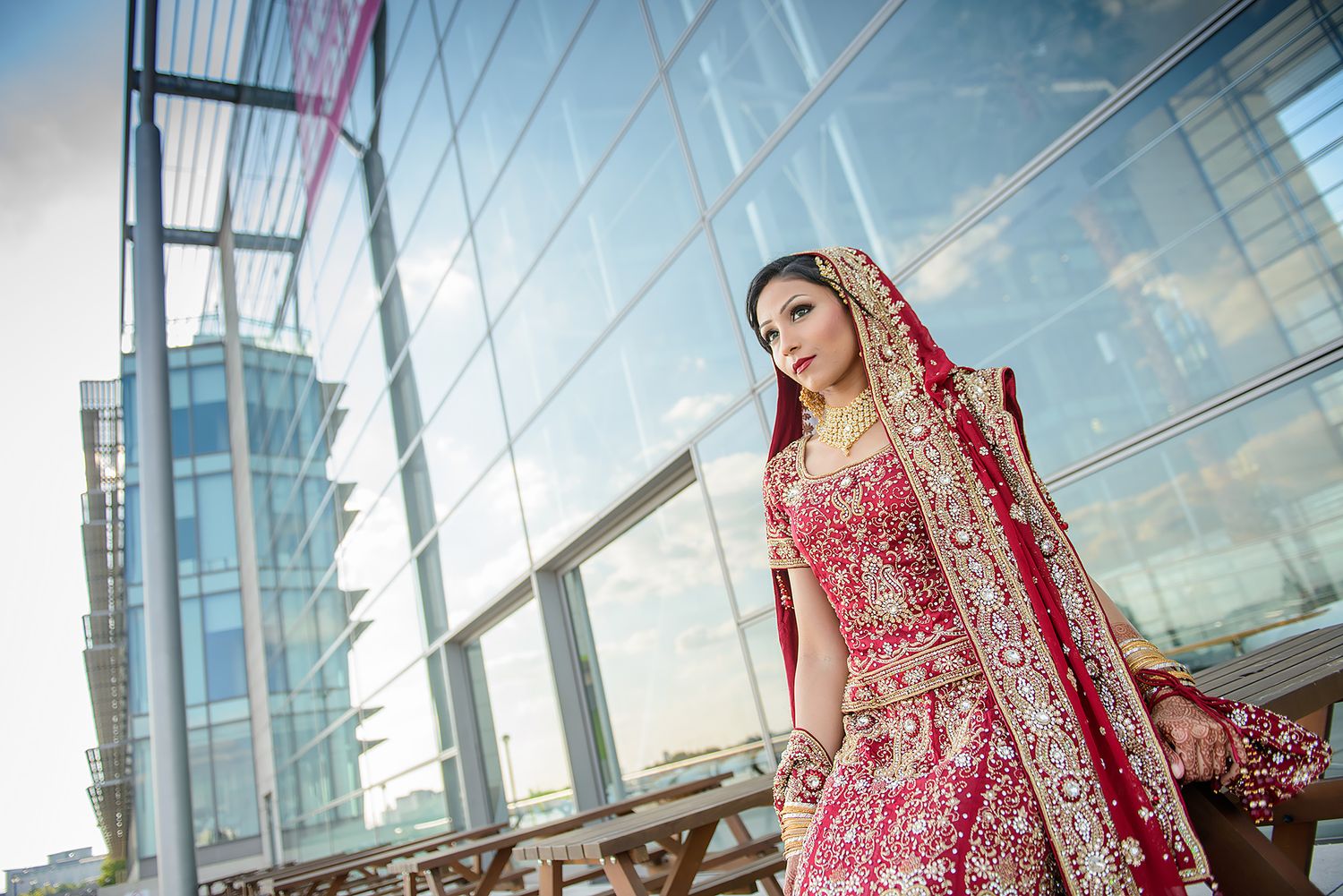 Bride in red lehenga poses against modern architecture with glass windows and city views.