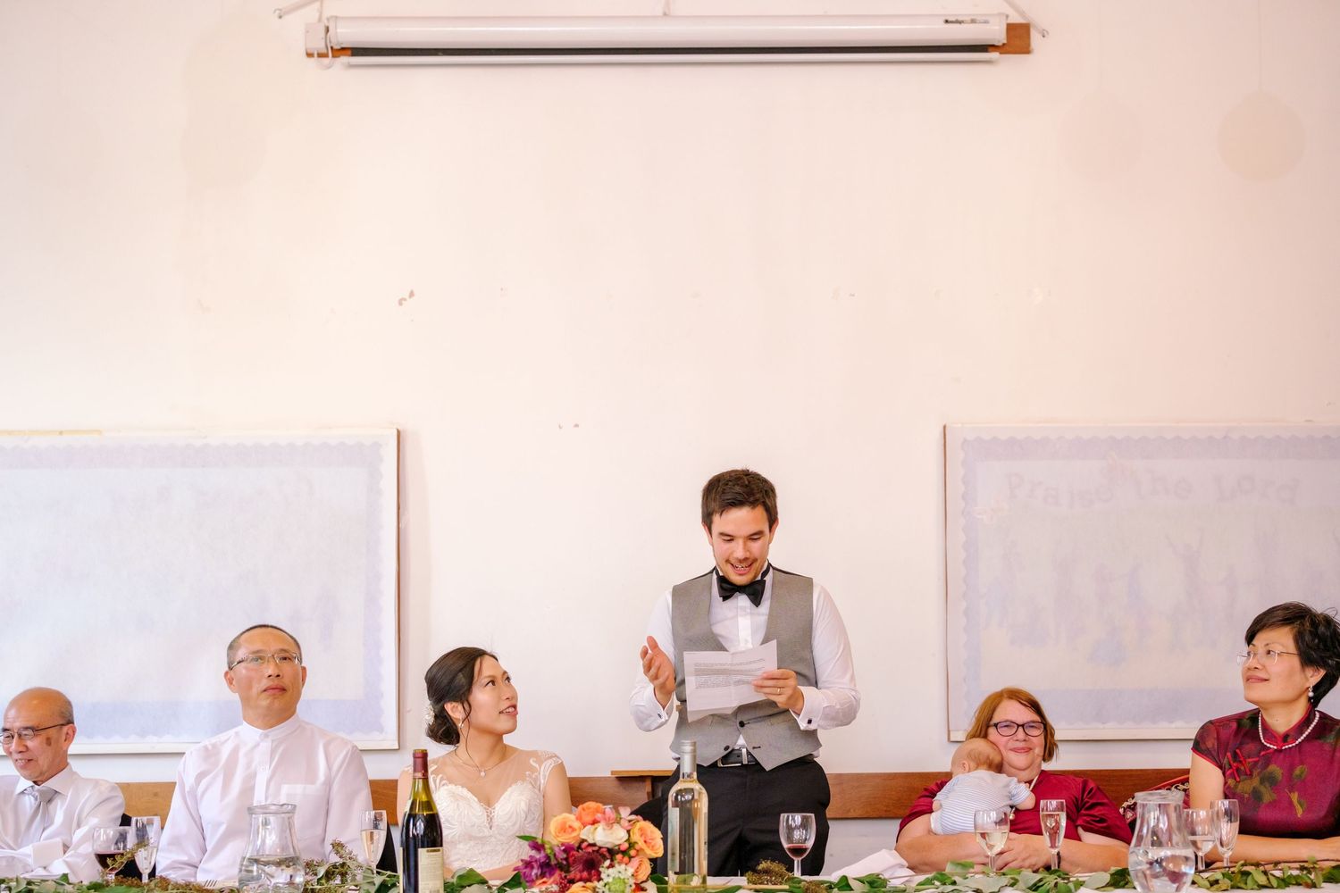 Wedding reception scene with guests seated at tables while someone gives a speech during dinner.