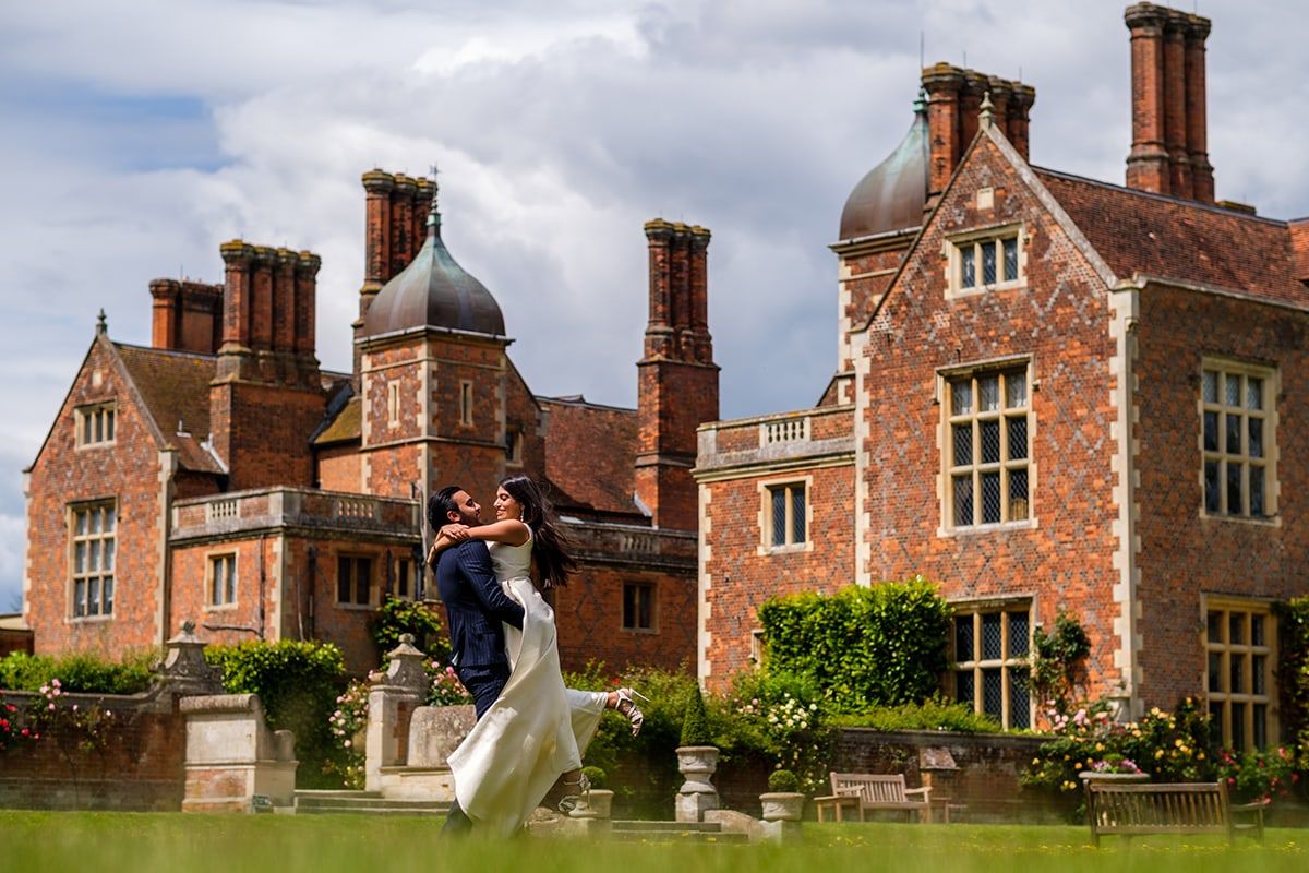Joyful wedding leap on the lawn of a historic Tudor-style mansion with distinctive chimneys.