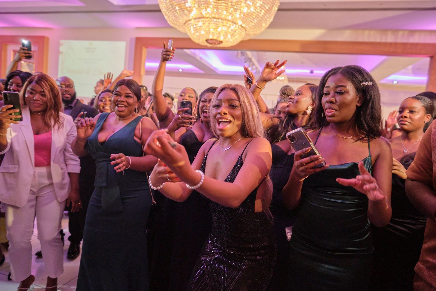 Wedding reception dance party with purple mood lighting and a large decorative chandelier overhead.