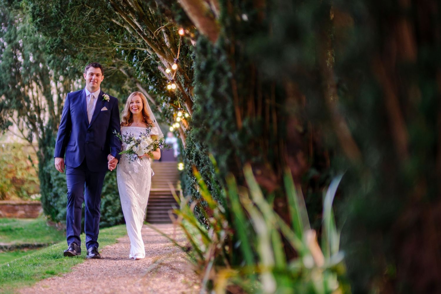 Couple walking along a tree-lined path during their outdoor wedding celebration.