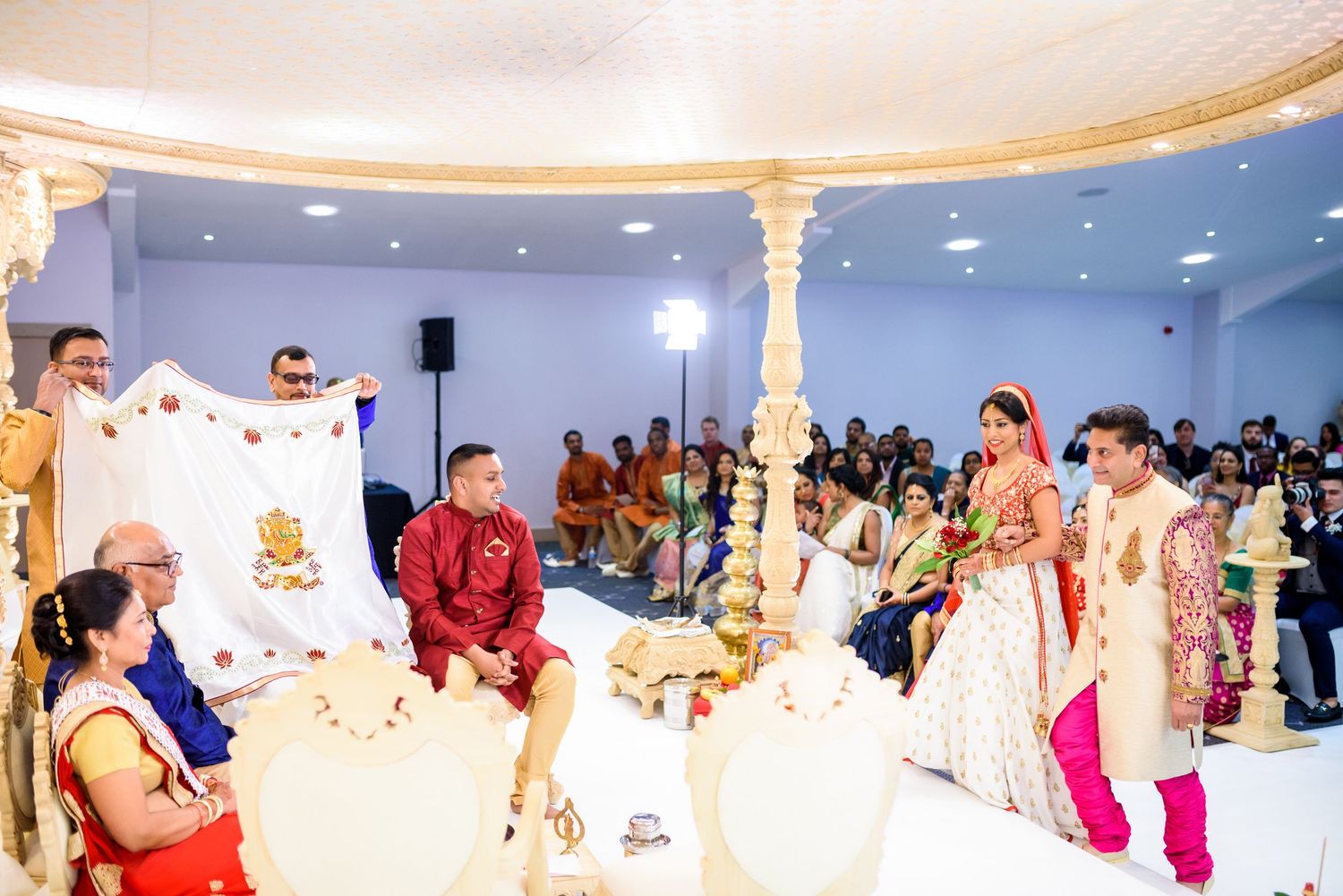 Traditional Indian wedding ceremony taking place under an ornate mandap with decorative pillars and gathered guests.