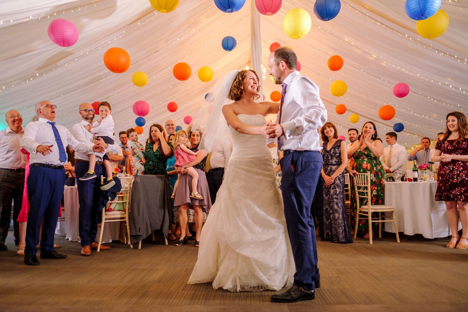 A wedding reception features colorful paper lanterns hanging from the ceiling as a couple dances on the floor.