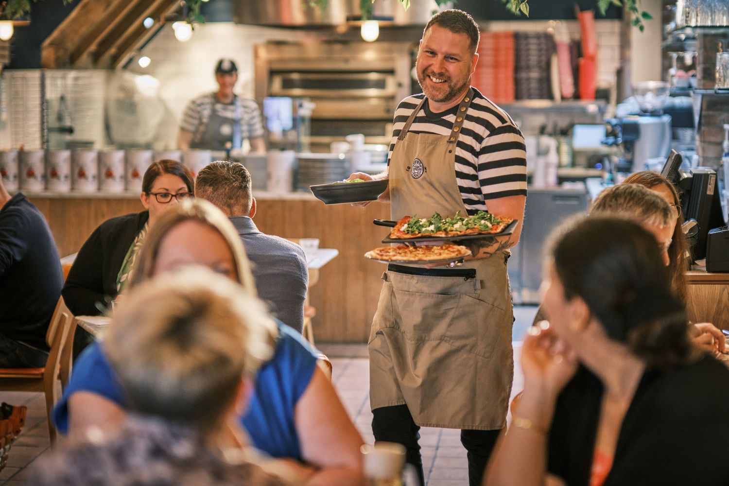 Restaurant server in striped shirt and apron delivering food to dining customers.