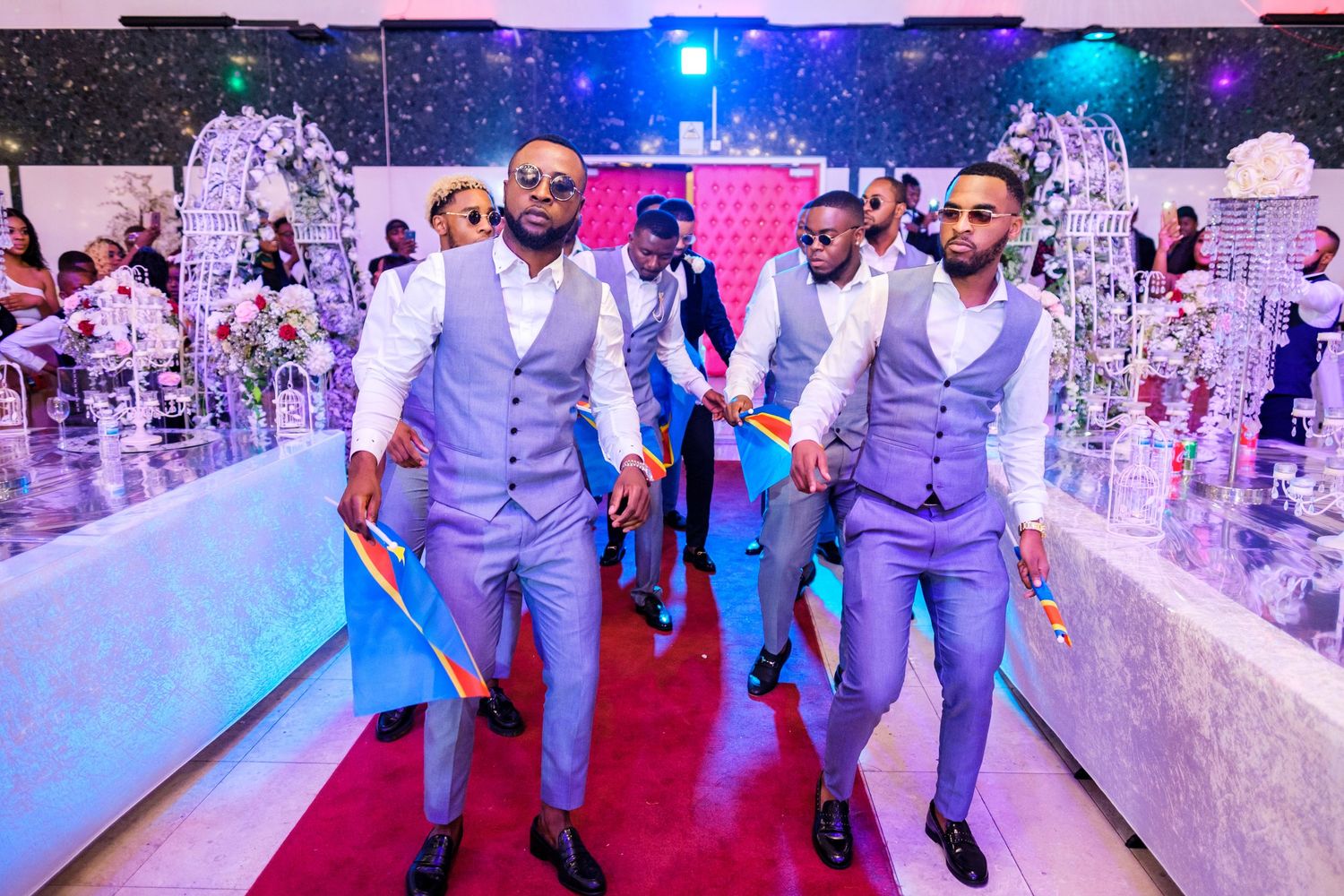 Wedding party members in light blue suits dance on a red carpet under vibrant lighting.