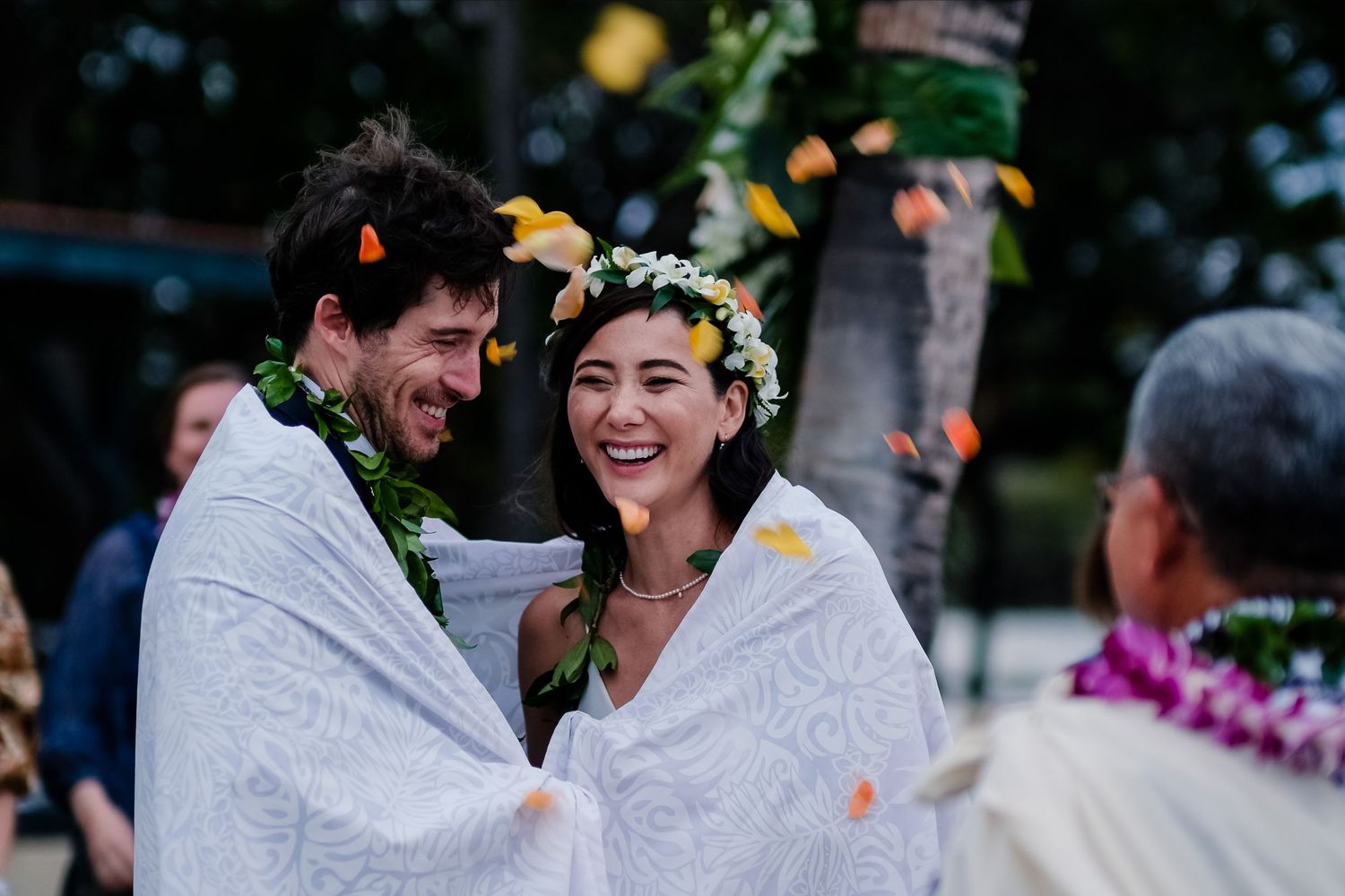 Happy couple sharing laughter during an outdoor celebration with flower petals falling.
